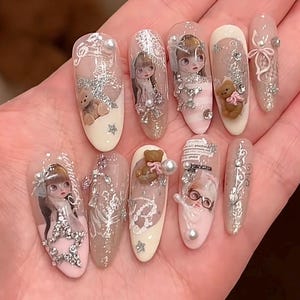 May include: A set of ten almond-shaped press-on nails featuring intricate designs including miniature 3D teddy bears, portraits of characters with large eyes, silver glitter accents, white lace patterns, small pearls, and clear rhinestones.