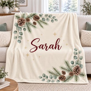 May include: A cream-colored fleece throw blanket featuring a printed design of pine branches, brown pinecones, and green eucalyptus leaves in the top-left and bottom-right corners. The name Sarah is printed in a dark red script in the center.