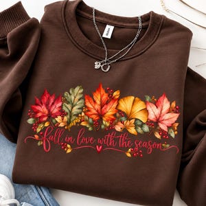 May include: A dark brown crewneck sweatshirt featuring a colorful graphic of autumn leaves, including red maple and yellow ginkgo foliage, with red berries and the text "fall in love with the season" written in a red cursive script font.