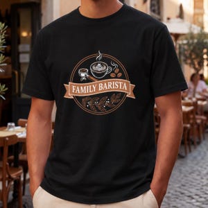 May include: A black short-sleeved t-shirt featuring a circular graphic with the text FAMILY BARISTA on a brown ribbon. The design includes a coffee cup with latte art, a portafilter, coffee beans, and a branch with leaves in white and brown.