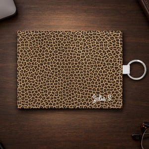 Puede incluir: Un tarjetero rectangular con un estampado de leopardo en tonos marrones y negros. El artículo incluye un llavero de metal plateado unido a una pestaña blanca en el lado derecho. El nombre Julia S. está impreso en blanco en la esquina inferior.