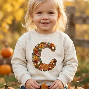 Könnte beinhalten: Ein cremefarbenes Langarm-Sweatshirt mit einem großen Buchstaben C als Grafik auf der Brust. Der Buchstabe besteht aus bunten Herbstblumen, Blättern und Eicheln, mit einer kleinen orangefarbenen Katze, die sich unten im Design zusammenrollt.
