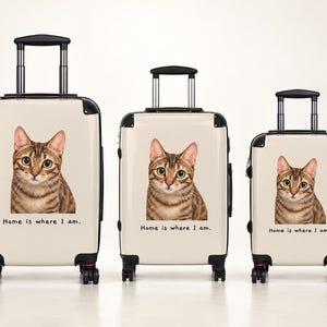 Könnte beinhalten: Drei cremefarbene Rollkoffer in verschiedenen Größen, jeweils mit einer braunen getigerten Katze und dem aufgedruckten Text Home is where I am. Die Gepäckstücke haben schwarze Eckschützer, schwarze Griffe und schwarze Rollen.