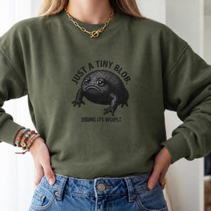 Könnte beinhalten: Ein olivgrünes Sweatshirt mit Rundhalsausschnitt und einer Grafik einer dunklen, runden Kröte mit großen Augen. Über der Kröte steht der Text "JUST A TINY BLOB." und unter der Kröte steht "DOING ITS WORST." Das Sweatshirt hat lange Ärmel und gerippte Bündchen.