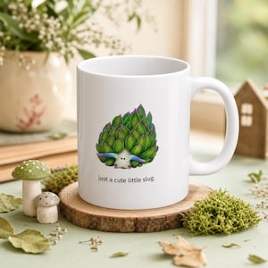 May include: A white ceramic mug featuring a graphic of a small slug covered in green succulent leaves with purple-tipped antennae. Below the illustration, the text reads, "just a cute little slug." The mug has a standard handle and a smooth finish.