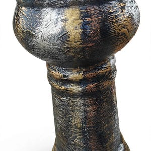 Könnte beinhalten: Eine dekorative Sockelvase mit einem breiten, gewellten Rand und einem zylindrischen Fuß. Die Oberfläche weist ein strukturiertes, abgenutztes Finish in den Farben Schwarz, Metallic-Gold und Silber auf, das mit sichtbaren Pinselstrichen eine verwitterte Optik erzeugt.