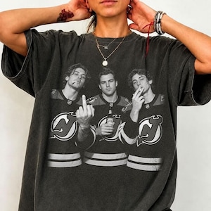 Może przedstawiać: Oversizowa koszulka w kolorze ciemnoszarym z czarno-białym nadrukiem graficznym przedstawiającym trzech hokeistów w koszulkach New Jersey Devils. Środkowy zawodnik trzyma butelkę, a gracze po bokach palą papierosy.