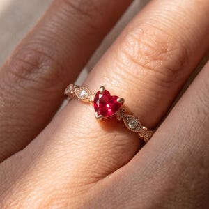 Può includere: Un anello in oro con una pietra preziosa rossa centrale a forma di cuore. La fascia è decorata con piccole pietre trasparenti incastonate in un motivo metallico che si estende lungo i lati dell'anello, creando un aspetto delicato e complesso.