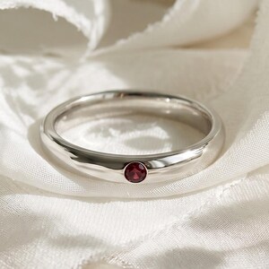 May include: A polished silver-toned band ring featuring a single, small, round, deep red gemstone set flush into the metal. The ring is positioned centrally, surrounded by soft white fabric ribbons and delicate white baby's breath flowers.