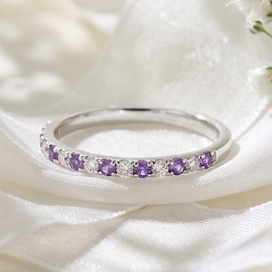 Op de afbeelding: Een zilverkleurige ring met een afwisselend patroon van ronde paarse amethisten en heldere witte diamanten. De ring is gezet in een gepolijste metalen band en toont een delicaat en symmetrisch ontwerp met meerdere edelstenen.