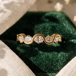 Può includere: Un anello in oro con un design ondulato continuo che sostiene cinque pietre chiare a taglio rotondo. Il metallo ha una finitura lucida e le pietre sono incastonate in fila lungo il centro dell'anello, creando un aspetto fluido e collegato.