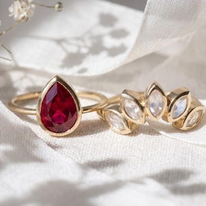 May include: A gold ring featuring a pear-shaped red gemstone in a bezel setting, positioned next to a curved gold band set with five marquise-cut iridescent white gemstones. Both rings are displayed on a textured white fabric surface.