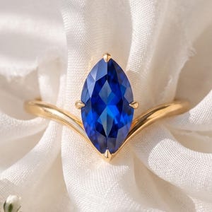 May include: A gold ring featuring a marquise-cut deep blue gemstone set in a V-shaped band. The stone is held by four prongs, and the band has a smooth, polished finish. The ring is positioned centrally, showcasing the vibrant blue color of the stone.