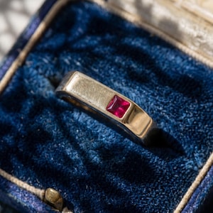 Peut inclure: Une bague en or avec un rubis rouge profond de taille carrée serti à fleur dans la surface métallique. La bague est présentée à l'intérieur d'une boîte à bijoux doublée de velours bleu foncé avec une bordure intérieure de couleur crème.
