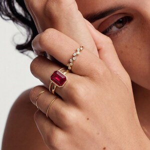 Puede incluir: Una colección de anillos de oro en los dedos, incluyendo un anillo prominente con una gran piedra roja rectangular en un engaste de oro, una banda delicada con pequeñas piedras claras redondas y varios anillos de oro finos, lisos y trenzados.