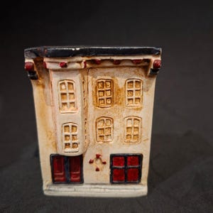 Può includere: Una statuetta di edificio in miniatura in ceramica con una facciata color crema e dettagli marroni usurati. La struttura presenta una finestra a bovindo sporgente, diverse finestre a griglia e due porte rosse con cornici scure al piano terra.