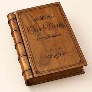 May include: A rectangular wooden book-shaped box with a dark brown finish. The cover features laser-engraved text reading Our Vows, Emma & James, and EST. JUNE 18, 2026, surrounded by decorative scrollwork borders and a spine with raised bands.