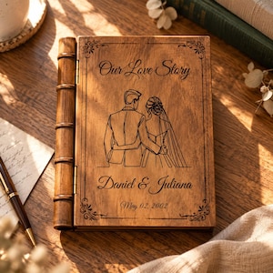 May include: A rectangular wooden book with a hinged spine, featuring a laser-engraved line art illustration of a couple, the text "Our Love Story," the names "Daniel & Juliana," and the date "May 02, 2002" framed by decorative corner flourishes.