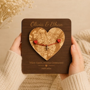 Può includere: Una targa quadrata in legno scuro con i nomi incisi Olivia & Ethan in alto. Una mappa in sughero a forma di cuore è centrata con due spilli a cuore rossi collegati da un filo rosso e un piccolo aereo in metallo. Il testo recita Miles Apart, Always Connected e 3459 miles.