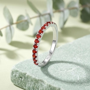 Könnte beinhalten: Ein silberner Ring mit einer Reihe kleiner, runder, leuchtend roter Edelsteine, die entlang der oberen Hälfte gefasst sind. Die Innenseite des Rings ist deutlich mit dem Stempel 925 versehen, was auf Sterlingsilber als Material hinweist.