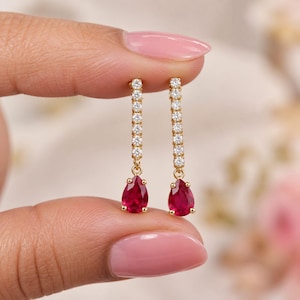 May include: A pair of gold drop earrings featuring a vertical row of small, clear, round-cut stones leading to a single, vibrant, pear-shaped red gemstone at the base. The earrings are crafted with a polished gold metal setting and secure clasps.