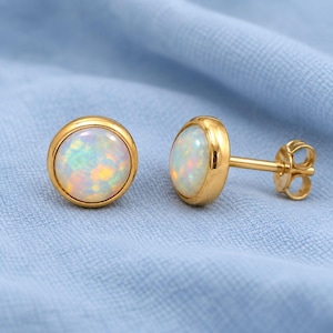 May include: A pair of round stud earrings featuring polished gold-tone settings and smooth, iridescent white opal cabochons. One earring is shown from the front, while the other is displayed from the side, revealing the post and butterfly back closure.