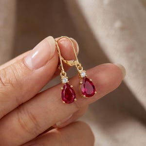Puede incluir: Un par de pendientes de oro con cierre de palanca, que presentan una piedra preciosa de color rojo intenso en forma de lágrima suspendida bajo una pequeña piedra clara de talla redonda. El engaste es de oro pulido con estilo colgante.