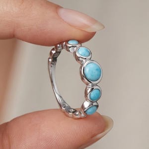 Könnte beinhalten: Ein Silberring mit fünf runden, hellblauen Larimar-Steinen in einer Reihe. Die Steine variieren in der Größe, wobei der größte Stein in der Mitte sitzt, flankiert von zwei kleineren Steinen auf jeder Seite, eingefasst in ein Silberband.