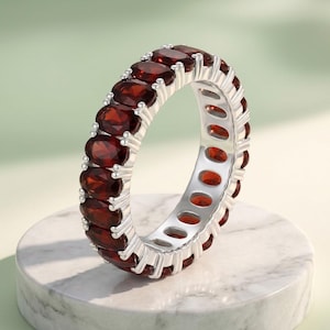 Op de afbeelding: Een eternity ring met een doorlopende rij ovaal geslepen, dieprode granaat edelstenen in een gepolijste, zilverkleurige metalen zetting. De ring toont een reeks stenen in klauwzetting die de band volledig en uniform omsluiten.