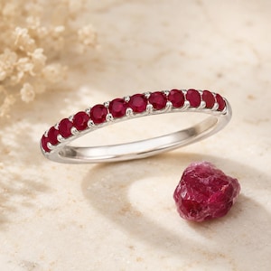 May include: A silver band ring featuring a row of eleven round-cut, deep red rubies set in a shared prong setting. Beside the ring sits a single, raw, unpolished deep red ruby gemstone with a jagged, natural texture and crystalline surface.