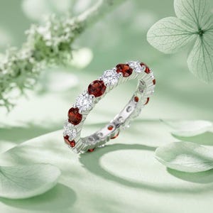 Op de afbeelding: Een zilveren eeuwigheidsring met een afwisselend patroon van rondgeslepen dieprode granaatedelstenen en heldere witte diamanten. De ring is vervaardigd met een gepolijste metalen zetting die elke steen stevig vasthoudt in een cirkelvormig ontwerp.
