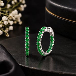 May include: A pair of silver hoop earrings featuring a continuous row of round-cut vibrant green gemstones set along the front-facing edge. The earrings have a hinged closure mechanism and a polished metallic finish, showcasing a circular design.