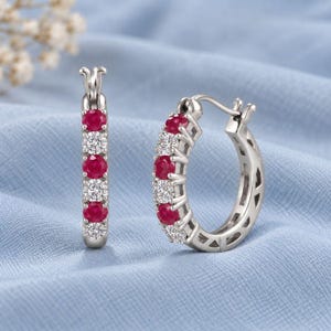 May include: A pair of silver-toned hoop earrings featuring an alternating pattern of round-cut red rubies and clear white stones. The earrings have a hinged closure mechanism and a polished metal finish, showcasing a symmetrical design.