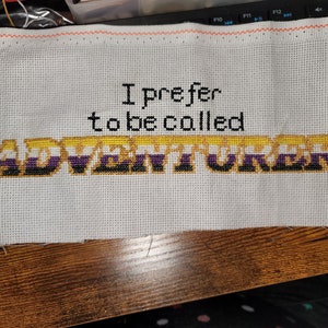 Puede incluir: Bordado de punto de cruz en tela blanca con el texto "I prefer to be called ADVENTURER" en amarillo, negro y morado.
