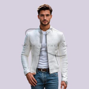 Puede incluir: Una chaqueta de cuero blanca con un patrón texturizado de piel de cocodrilo, con cuello alzado, cremallera frontal completa y dos bolsillos laterales con cremallera. La chaqueta se lleva sobre una camiseta blanca lisa y vaqueros azules con cinturón marrón.