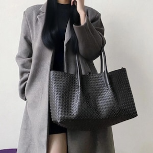Puede incluir: Un bolso tote negro grande con un intrincado patrón de cuero trenzado, dos asas superiores resistentes y una silueta rectangular espaciosa. El bolso está construido con una textura trenzada uniforme en todo el exterior y asas de cuero liso.