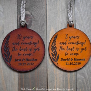 May include: Two round leather ornaments with a brown and orange color scheme. The ornaments are engraved with the text "10 years and counting! The best is yet to come... Josh & Heather 10.27.2012" and "3 years and counting! The best is yet to come... David & Hannah 11.10.2019".