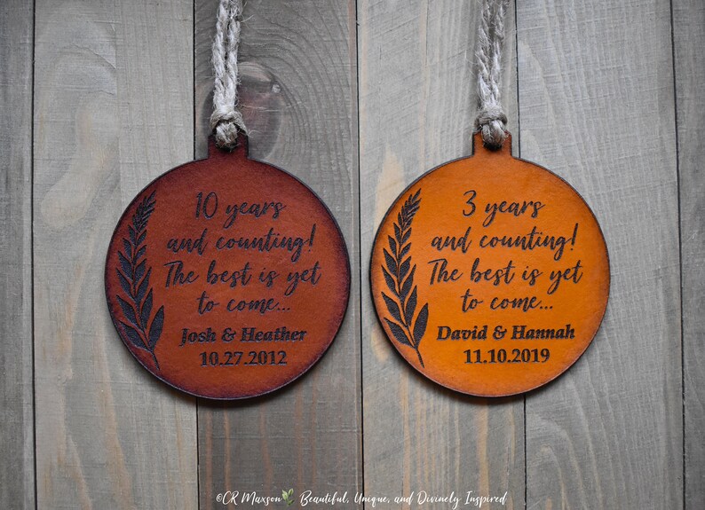 May include: Two round leather ornaments with a brown and orange color scheme. The ornaments are engraved with the text "10 years and counting! The best is yet to come... Josh & Heather 10.27.2012" and "3 years and counting! The best is yet to come... David & Hannah 11.10.2019".