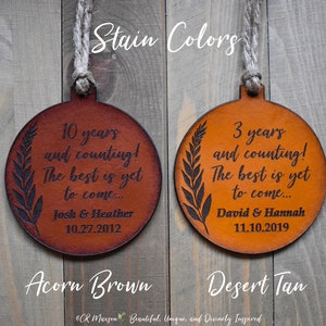 May include: Two leather ornaments with a leaf design and personalized text. The ornament on the left is acorn brown and says "10 years and counting! The best is yet to come... Josh & Heather 10.27.2012". The ornament on the right is desert tan and says "3 years and counting! The best is yet to come... David & Hannah 11.10.2019".