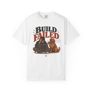 Puede incluir: Una camiseta blanca de manga corta con un gráfico de una persona sentada con un portátil y un perro golden retriever. El texto BUILD FAILED aparece sobre la persona y Ball still works en un bocadillo. Hay una huella en la parte inferior.