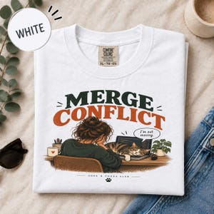 Peut inclure: Un t-shirt blanc avec une illustration représentant une personne assise à un bureau avec un ordinateur portable et un chat allongé sur le clavier. Le texte indique "MERGE CONFLICT", "I'm not moving." dans une bulle et "CODE & CHAOS CLUB".