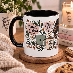 May include: A white ceramic mug with a black handle and interior, featuring a colorful graphic print of a laptop, a coffee mug, leaves, and hearts. The text reads Mama Codeur, I AM STRONG AF, SAME GIRL BIGGER GOALS, and Fueled by Coffee and Code.