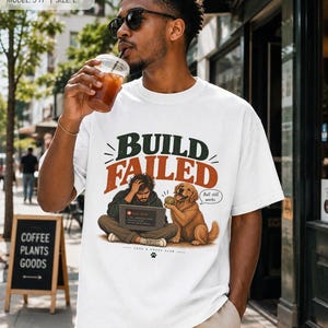 Könnte beinhalten: Ein weißes Kurzarm-T-Shirt mit einem Grafikdruck einer Person, die vor einem Laptop mit der Aufschrift BUILD FAILED sitzt, und einem Golden Retriever, der einen Tennisball hält, mit einer Sprechblase, die Ball still works sagt.