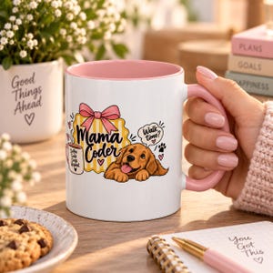 Op de afbeelding: Een witte keramische mok met een roze binnenkant en handvat, voorzien van een afbeelding van een golden retriever pup, een roze strik en de tekst Mama Coder, plus een kleine mokillustratie met Coffee Code Dogs Repeat en tekst Walk time.