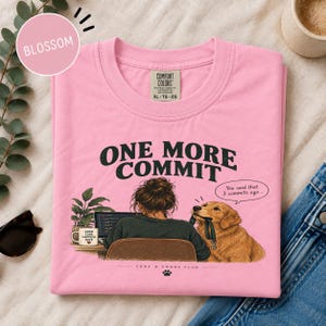 May include: A pink t-shirt featuring a graphic of a person sitting at a desk with a laptop, a potted plant, and a coffee mug labeled "GOOD CODE HAPPIER DAYS." A golden retriever sits next to them with a leash in its mouth. Text reads "ONE MORE COMMIT" and "You said that 3 commits ago..." and "CODE & CHAOS CLUB."