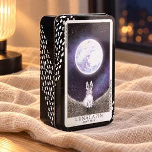 Op de afbeelding: Een zwarte rechthoekige metalen doos voor het Lunalapin Tarot-deck. De voorzijde toont een witomrande illustratie van een konijn dat op een heuvel met gras zit onder een grote volle maan tegen een donkere sterrenhemel. De zijkanten zijn versierd met kleine witte konijnensilhouetten.