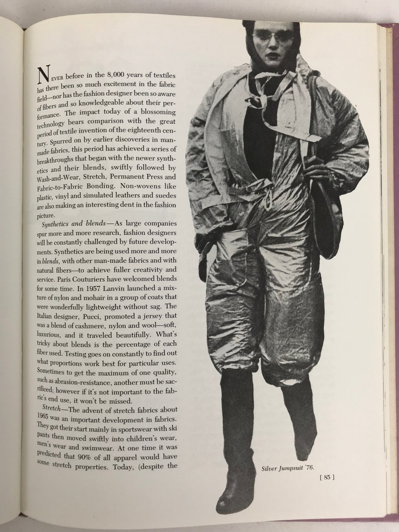 May include: A black and white photograph of a person wearing a silver jumpsuit. The person is standing with their legs slightly apart and their arms at their sides. They are wearing a silver jumpsuit with a zipper down the front and a silver belt. The person is looking at the camera. The text "Silver Jumpsuit 76." is printed in the bottom right corner of the image.