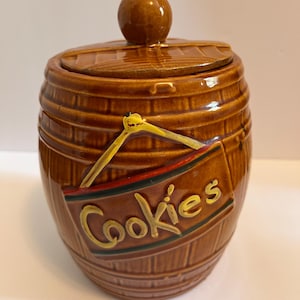Pode incluir: Pote de biscoitos de cerâmica marrom em forma de barril, com tampa e botão redondo. O pote tem uma placa que diz "Cookies" em letras amarelas.