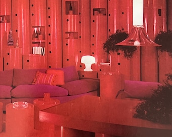 May include: A living room interior featuring a wall of tall, cylindrical red columns with integrated shelving units. A purple sofa with orange pillows sits in front of the columns. A white mushroom-shaped lamp and a clear pendant light are visible.