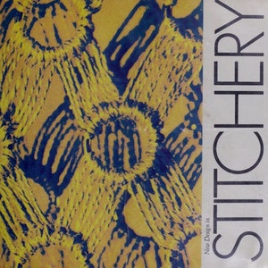 Pode incluir: Uma capa de livro com o título "Stitchery" em letras brancas grandes sobre um fundo branco. A capa apresenta um close-up de um design abstrato amarelo e azul.